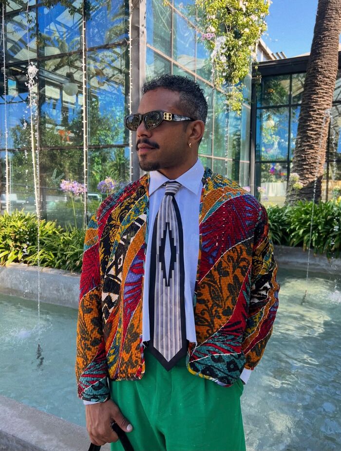 Yoel de tres cuartos, con lentes de sol, camisa blanca y la corbata
                  suelta, bajo una chaqueta de patchwork en rojos, naranjas, amarillos y
                  turquesa: piezas distintas cosidas en una sola. Detrás, una fuente y un
                  invernadero de vidrio a pleno sol.